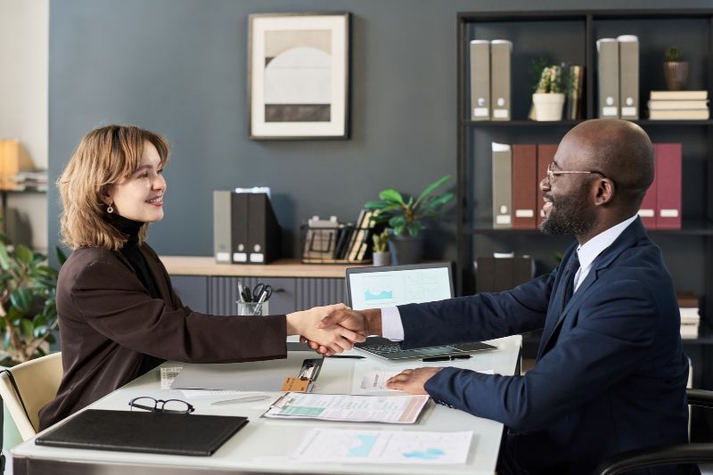 Two professionals shaking hands across a desk during a business meeting in an office.