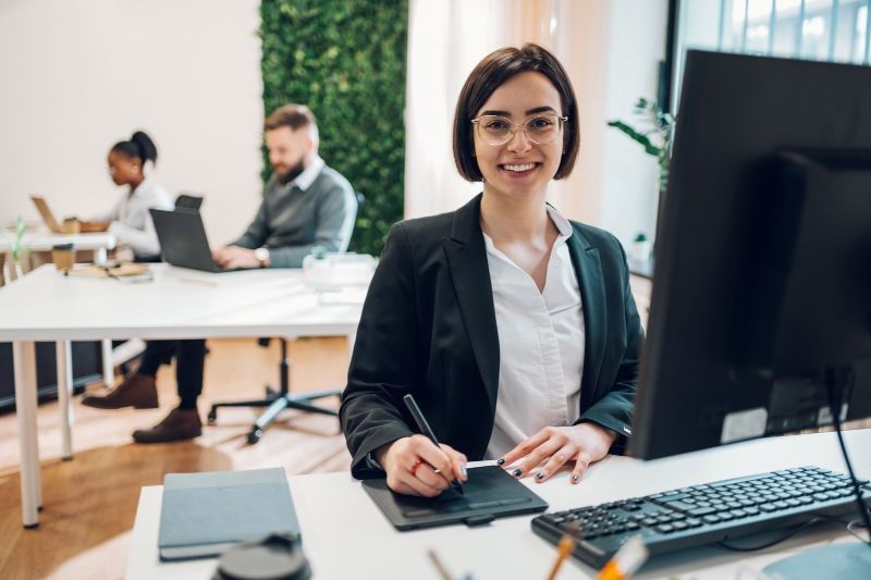 Professional woman working at a computer in a modern office with colleagues in the background.