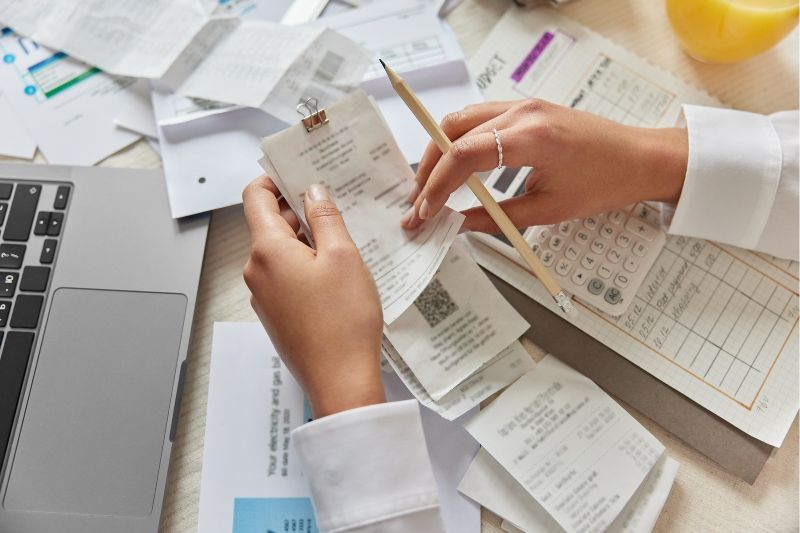 Person reviewing receipts and calculating expenses with a laptop, calculator, and financial documents.