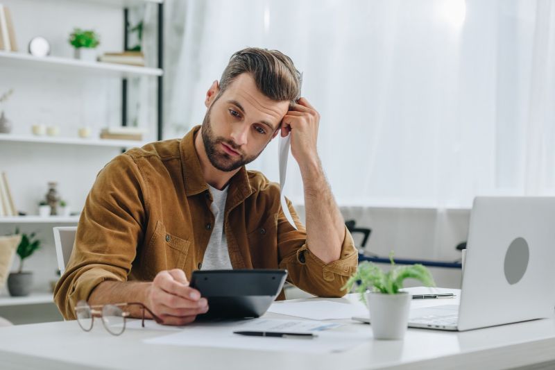 Freelancer reviewing expenses with a calculator and laptop in a home office.